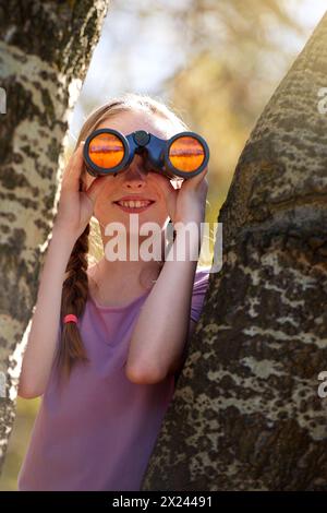 Albero, ragazza e binocolo per safari, ricerca e ricerca di animali in vacanza. Sorridi, divertiti e cerca di osservare gli uccelli selvatici o la fauna selvatica in natura Foto Stock