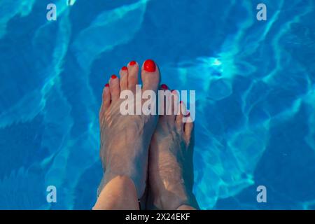 Piedini femminili con unghie rosse lucide immerse in una piscina d'acqua blu Foto Stock