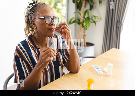 Donna afroamericana anziana a casa che tiene un bicchiere d'acqua, prende medicine Foto Stock