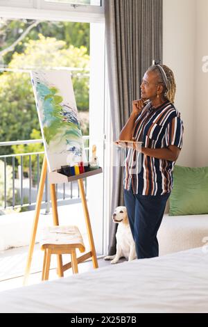 Una donna afroamericana anziana con trecce grigie in camicia a righe guarda l'arte a casa Foto Stock