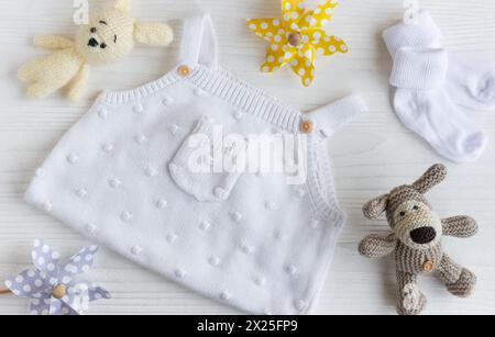 Set di abiti bianchi e accessori per neonato. Giocattoli a maglia, rompicapo a maglia, calze su sfondo bianco in legno. Concetto di baby shower. Piatto, Foto Stock