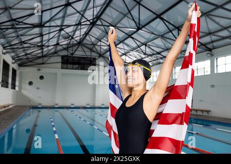 Una giovane nuotatrice birazziale che regge una bandiera americana si trova a bordo piscina all'interno, spazio per copiare Foto Stock