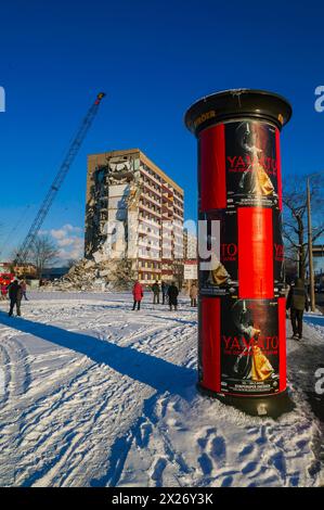 Le persone passano davanti a un pilastro pubblicitario mentre un edificio viene demolito sullo sfondo Foto Stock