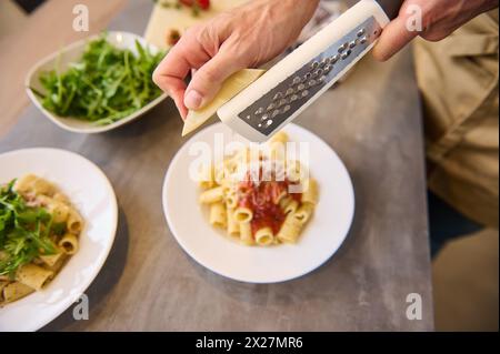 Direttamente sopra le mani dello chef usando la grattugia, grattugiare il formaggio sulla pasta appena cotta con salsa di pomodoro, condire e riempire il pasto prima di servirlo. Foto Stock