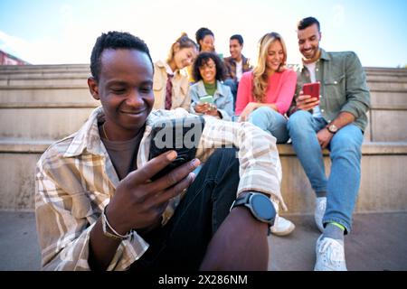 Un ragazzo nero sorridente che usa lo smartphone. Uomo afroamericano che tiene la cella all'aperto con gli amici Foto Stock