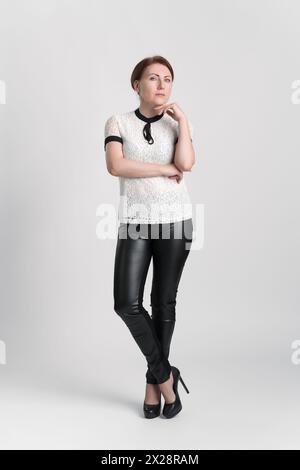 Donna d'affari sicura che guarda la fotocamera con una mano sollevata al mento, gambe in piedi incrociate alla caviglia su sfondo bianco. Donna d'affari Foto Stock