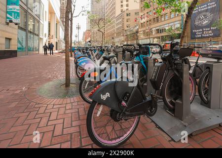 San Francisco, California, 8 aprile 2024. Le biciclette ancorate attendono i ciclisti su un marciapiede di San Francisco con la vita urbana sullo sfondo. Foto Stock