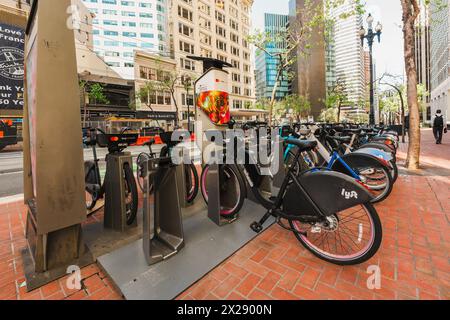 San Francisco, California, 8 aprile 2024. Le biciclette ancorate attendono i ciclisti su un marciapiede di San Francisco con la vita urbana sullo sfondo. Foto Stock