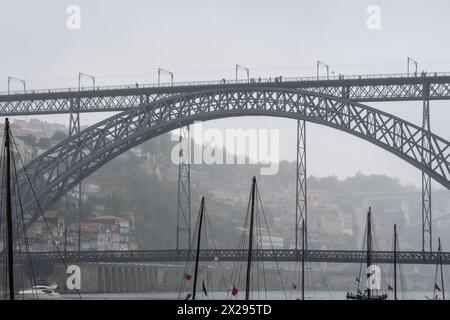 Alberi di barche rabelos in legno attraccati sul fiume Douro a Porto, sotto il ponte in acciaio Don Luis i sullo sfondo avvolti dalla nebbia su una pioggia grigia Foto Stock