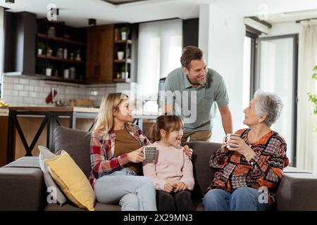 Una scena emozionante si svolge mentre una famiglia multigenerazionale si riunisce su un divano per presentare una torta di compleanno a una nonna adorata, creando ricordi indimenticabili Foto Stock