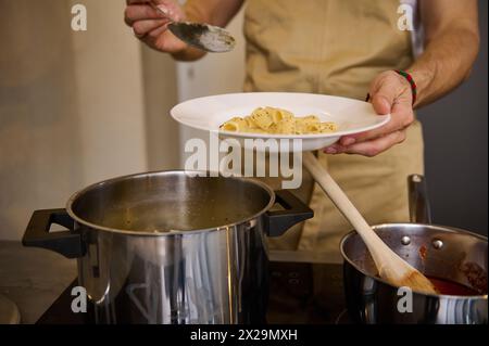 Primo piano, uno chef maschile che sta in piedi davanti a una stufa elettrica e mette la pasta appena cucinata su un piatto bianco Foto Stock
