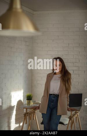 Giovane donna appoggiata a un tavolo con un portatile nelle vicinanze, inondata di luce naturale da una grande finestra. La scena suggerisce un lavoro produttivo Foto Stock