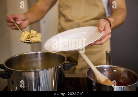 Primo piano, uno chef maschile che sta al piano cottura e mette penne di pasta appena cucinata su un piatto bianco. Concetto alimentare Foto Stock
