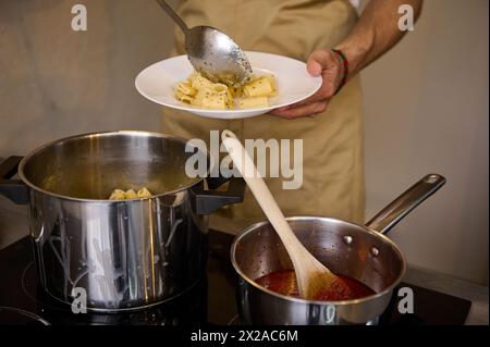 Le mani dello chef da vicino preparano pasta appena cucinata, mettono penne bollite su un piatto bianco, stando in piedi davanti alla stufa elettrica a induzione con la pentola piena Foto Stock