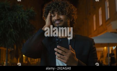 Giovane indiano arabo latino americano uomo che usa smartphone cellulare SMS chattare online strada all'aperto ragazzo che tiene la testa problema mal di testa Foto Stock