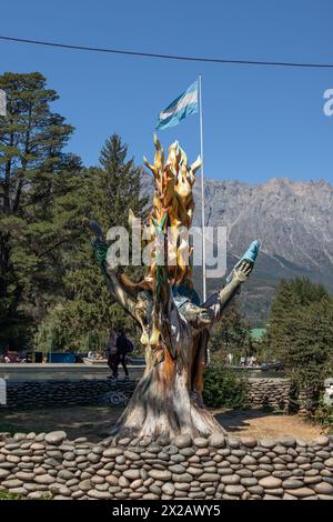 Scultura di alberi in una piazza con una bandiera argentina dietro e alberi e montagne come sfondo a El Bolsón Foto Stock