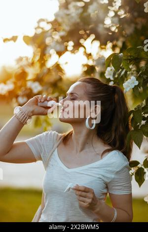 Ora legale. donna elegante in camicia bianca con spray nasale vicino all'albero fiorito. Foto Stock