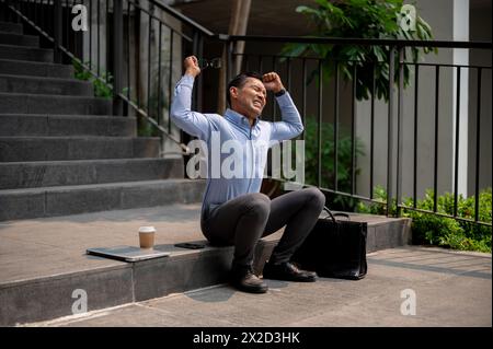 Un uomo d'affari asiatico arrabbiato e stressato si siede sui gradini all'aperto, piangendo, urlando, sperimentando stress o delusione, venendo licenziato dal suo lavoro. Foto Stock