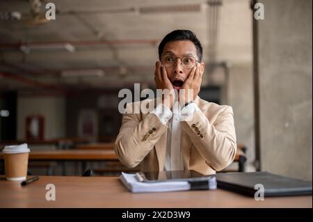 Un imprevisto uomo d'affari millenario asiatico sorpreso tiene la faccia sotto shock, la bocca spalancata e gli occhi spalancati, siede a un tavolo con le carte in un bullo Foto Stock