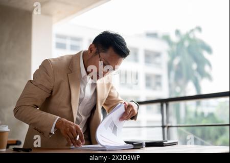 Un uomo d'affari asiatico serio e determinato che esamina i rapporti, cerca un documento, si concentra su un documento su un tavolo, lavora al t Foto Stock