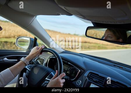 Vista interna di una donna che guida un'auto su una strada di campagna. Conducenti femminili mani sul volante Foto Stock