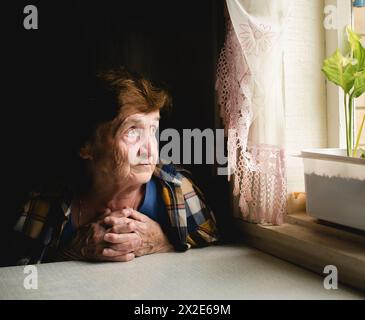 Una donna anziana siede accanto alla finestra, guardando fuori con un'espressione premurosa, persa nella contemplazione. Foto Stock