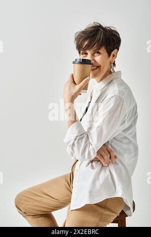 Un'elegante donna di mezza età con i capelli corti si siede su una sedia, tenendo in mano una tazza di caffè. Foto Stock