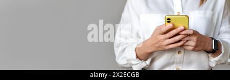 Donna d'affari irriconoscibile che indossa messaggi online in camicia bianca utilizzando il suo smartphone su sfondo grigio. Foto Stock