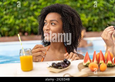 Giovane donna che sorseggia succhi di frutta a bordo piscina - gusta spuntini salutari - ambiente estivo rilassato, rinfresco naturale. Foto Stock