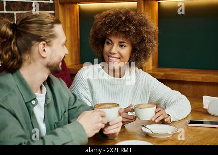 Una donna afroamericana e un uomo si siedono a un tavolo, gustando tazze di caffè in un moderno ambiente da caffè. Foto Stock