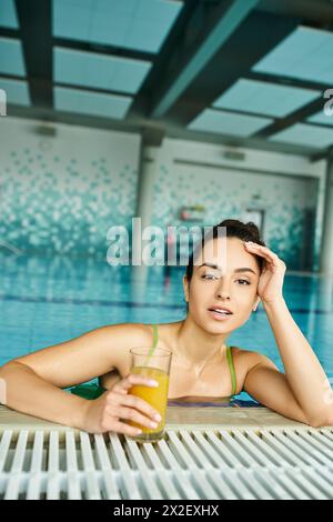 Una giovane donna bruna in costume da bagno gode di un bicchiere di succo d'arancia mentre si rilassa in una piscina termale interna. Foto Stock