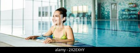 Una giovane donna bruna in un vivace bikini verde si rilassa in una tranquilla piscina interna spa. Foto Stock
