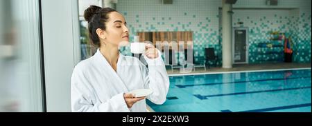 Una giovane donna bruna si gode una tranquilla mattinata sorseggiando un caffè in un lussuoso accappatoio presso una piscina spa interna. Foto Stock