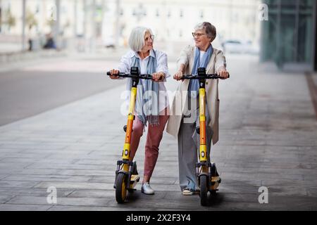 Le amiche mature e gioiose donne dai capelli grigi guidano gli scooter elettrici in città, sorridono e si divertono insieme. Foto Stock