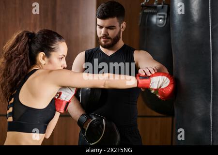 Un allenatore maschile si trova accanto a una donna sportiva bruna che indossa guanti da boxe, allenandosi attivamente in una palestra. Foto Stock