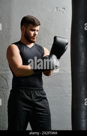 Un bell'uomo con la barba si trova accanto a una borsa da pugno in palestra, praticando tecniche di pugilato con concentrazione e determinazione. Foto Stock