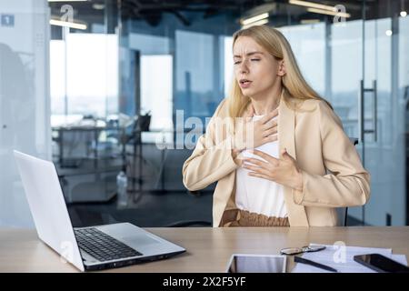 Una giovane donna d'affari sente un improvviso dolore toracico, respira alla scrivania dell'ufficio con laptop, occhiali e documenti in giro. Foto Stock