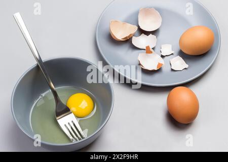 Uova rotte e forchetta in un recipiente grigio. Gusci di pollo su piatto grigio. Due uova marroni sul tavolo. Sfondo grigio. Vista dall'alto Foto Stock