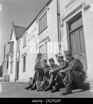 TRUPPE STATUNITENSI IN UN VILLAGGIO INGLESE: VITA DI TUTTI I GIORNI CON GLI AMERICANI A BURTON BRADSTOCK, DORSET, INGHILTERRA, Regno Unito, 1944 - quattro soldati americani siedono al sole sul marciapiede fuori dall'Anchor Inn a Burton Bradstock e chiacchierano con la ragazza locale Betty 'Freckles' Mackay. Secondo la didascalia originale, il soprannome "Freckles" è stato dato a Betty dal GIS, che l'hanno "resa una favorita del campo" Foto Stock