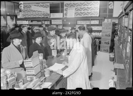 Uomini, donne e bambini fanno la fila al Selfridge's Grocery Department di Londra, 1942, durante lo shopping in tempo di guerra. Gli assistenti del negozio indossano cappotti bianchi e gli scaffali espongono vari prodotti alimentari, tra cui frumento tritato, oranjeast, spinaci di Smedley e chicchi d'orzo. Foto Stock