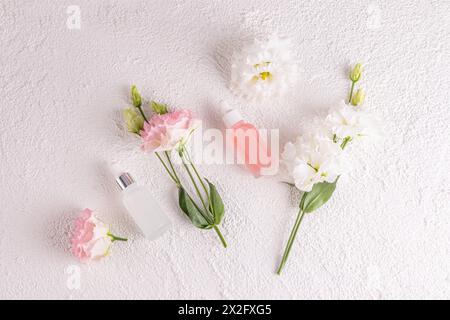 Due bottiglie cosmetiche opache con contagocce e un prodotto naturale per la cura della pelle per il viso su sfondo bianco tra delicati fiori primaverili. Posizione piatta. Foto Stock