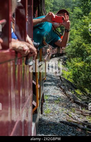 Uomo che si sporge dal finestrino di un vagone ferroviario all'aperto della Great Smoky Mountains Railroad per scattare una foto durante un'escursione da Bryson City, nord Foto Stock