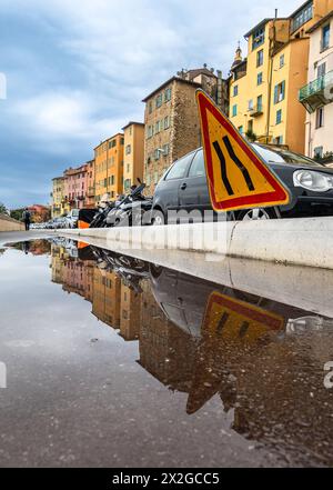 I segnali stradali che restringono la strada, le automobili e le case a Mentone si riflettono in una pozzanghera dopo la pioggia Foto Stock