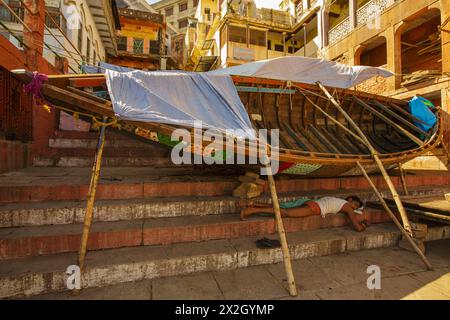 Workman che dormiva sotto una barca si fermò su Meer Ghat per riparazioni a Varanasi, in India Foto Stock