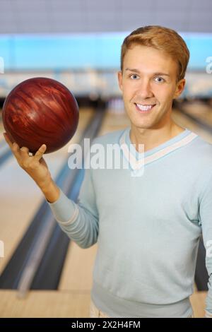 Un uomo bellissimo che indossa un poluver blu tiene la palla rossa nel club di bowling; profondità di campo bassa Foto Stock