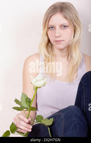 La ragazza con la rosa bianca si siede con calma e un po' malinconica con la schiena premuta contro il muro. Foto Stock