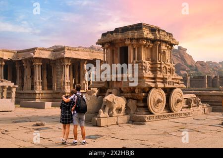 Coppia di turisti goditi la vista dell'antico carro in pietra con altre rovine medievali al complesso del tempio Vijaya Vittala a Hampi, Karnataka, India Foto Stock