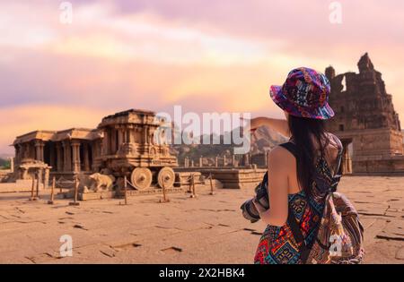 Giovani donne turistiche puntano verso l'antico carro di pietra di Hampi, Karnataka, India, al tramonto. Foto Stock