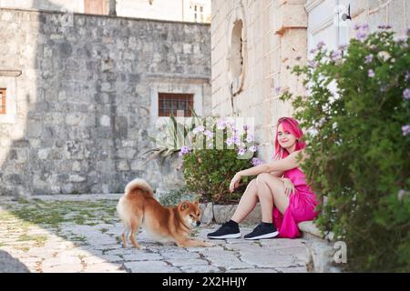 Una donna dai capelli rosa vivaci accarezza delicatamente un cane Shiba Inu in un pittoresco vicolo lastricato di pietra, un tenero momento di connessione. Foto Stock