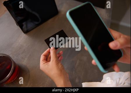 Le mani di una donna in primo piano tengono in mano una carta di credito nera con uno spazio pubblicitario per la copia del testo e utilizzano uno smartphone per lo shopping online, la ricezione o il pagamento Foto Stock